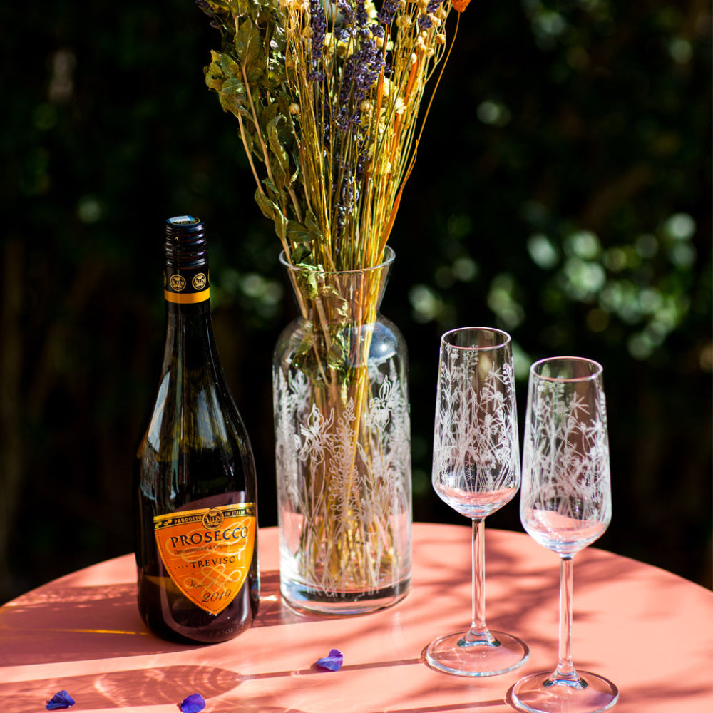 Prosecco bottle, vase with flowers, and two glasses on a table outdoors.