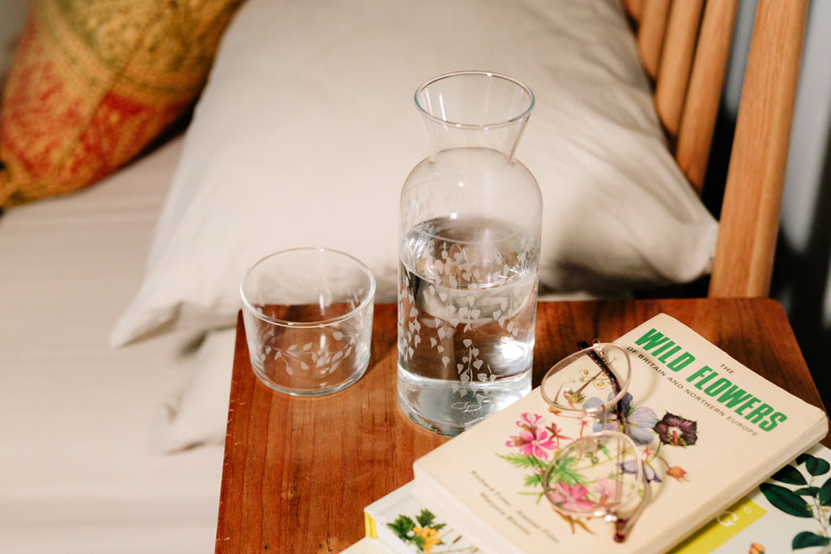 Clear glass vase and two glasses on a wooden table with a book titled 'Wild Flowers' in the background.