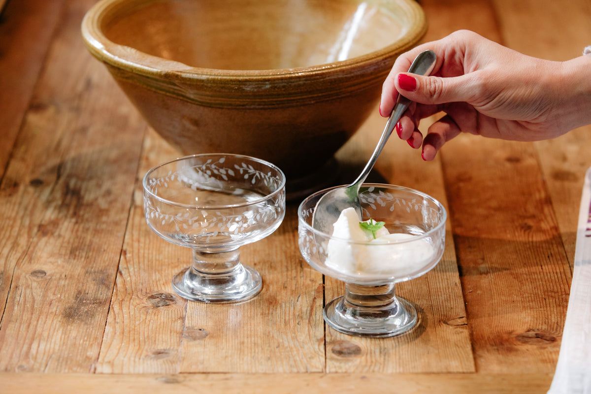 Person using a spoon to serve dessert into a glass bowl on a wooden table.