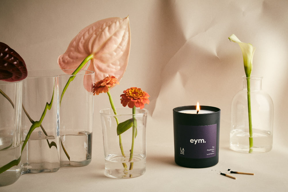 Candle with 'eym' branding next to flowers in clear vases on a light background