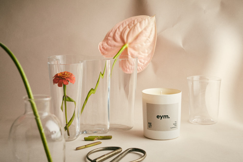 Candle with 'eym.' branding next to flowers and vases on a neutral background