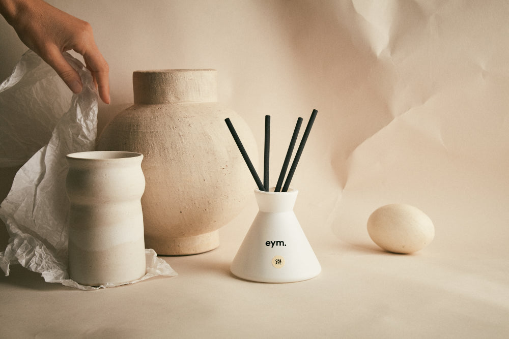 White diffuser with black sticks on a beige surface, surrounded by natural ceramic items.