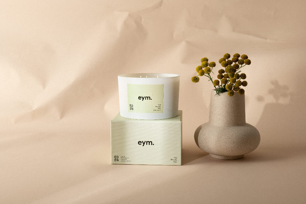 Three wick Candle called 'Eden' and packaging with 'eym.' branding on a beige background with a vase of flowers.