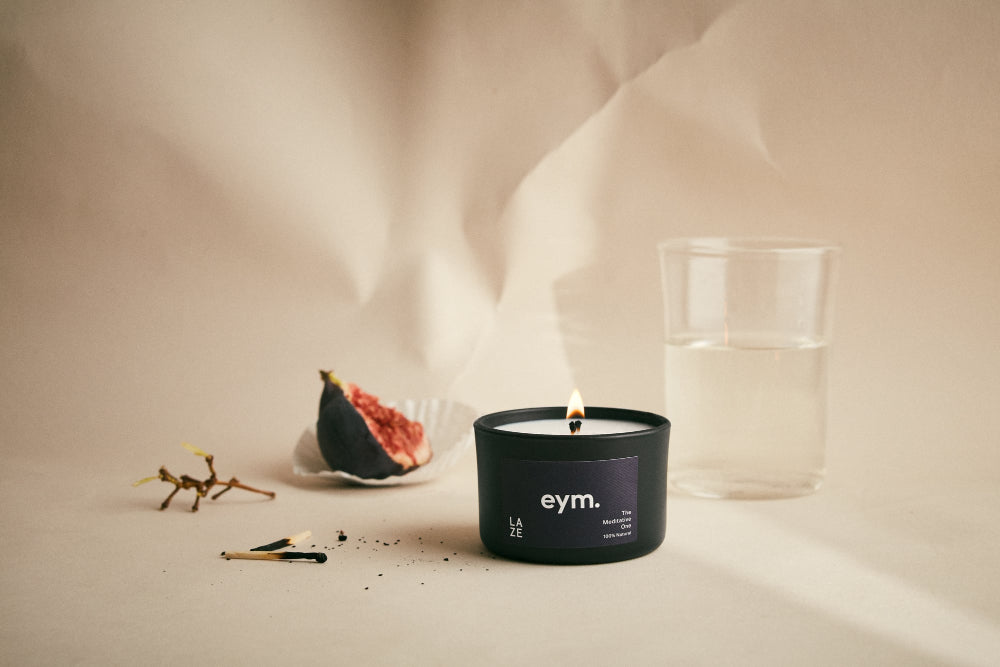 Candle with 'eym' branding on a neutral background with figs and a water vase in the background