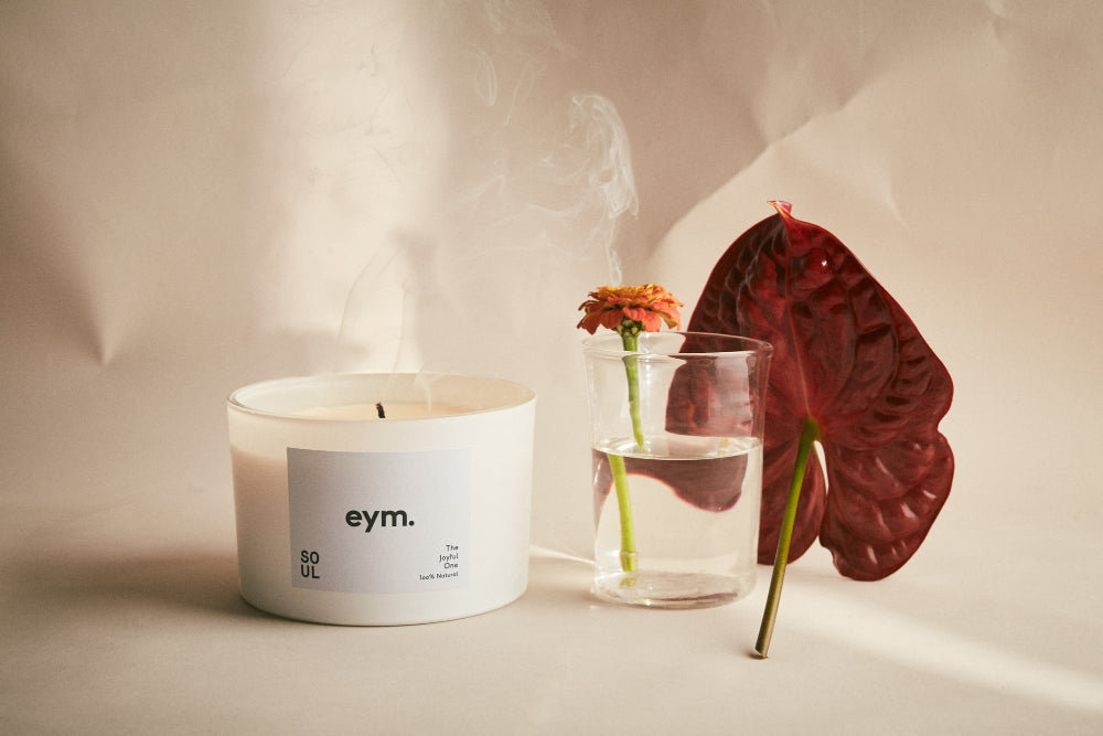 Candle with 'eym.' label next to a glass with flowers and a red leaf on a neutral background