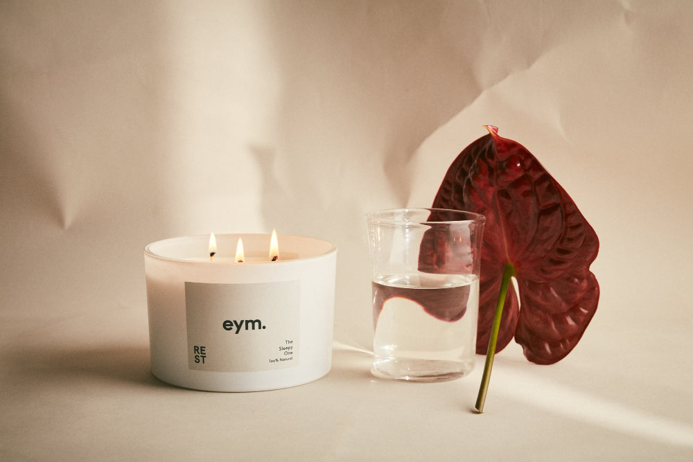 Candle with 'eym.' label next to a glass of water and a red leaf on a neutral background