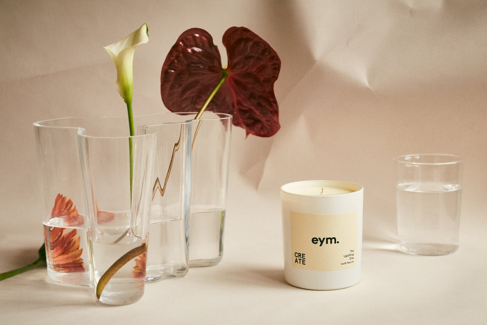 Candle with 'eym.' branding next to glass containers with flowers on a neutral background