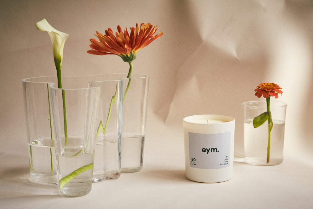 Candle called SOUL with 'eym' label surrounded by glass vases with flowers on a neutral background
