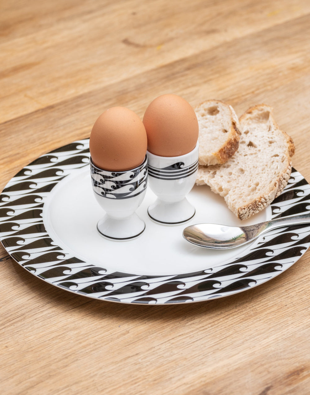 Two eggs in black and white striped egg cups on a decorative plate with bread and a spoon.