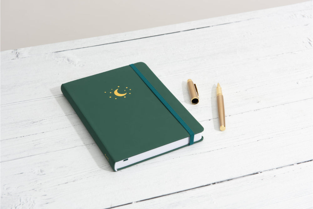 Forest Green Yop and Tom notebook with a gold moon and stars on a white wooden surface with two pens.