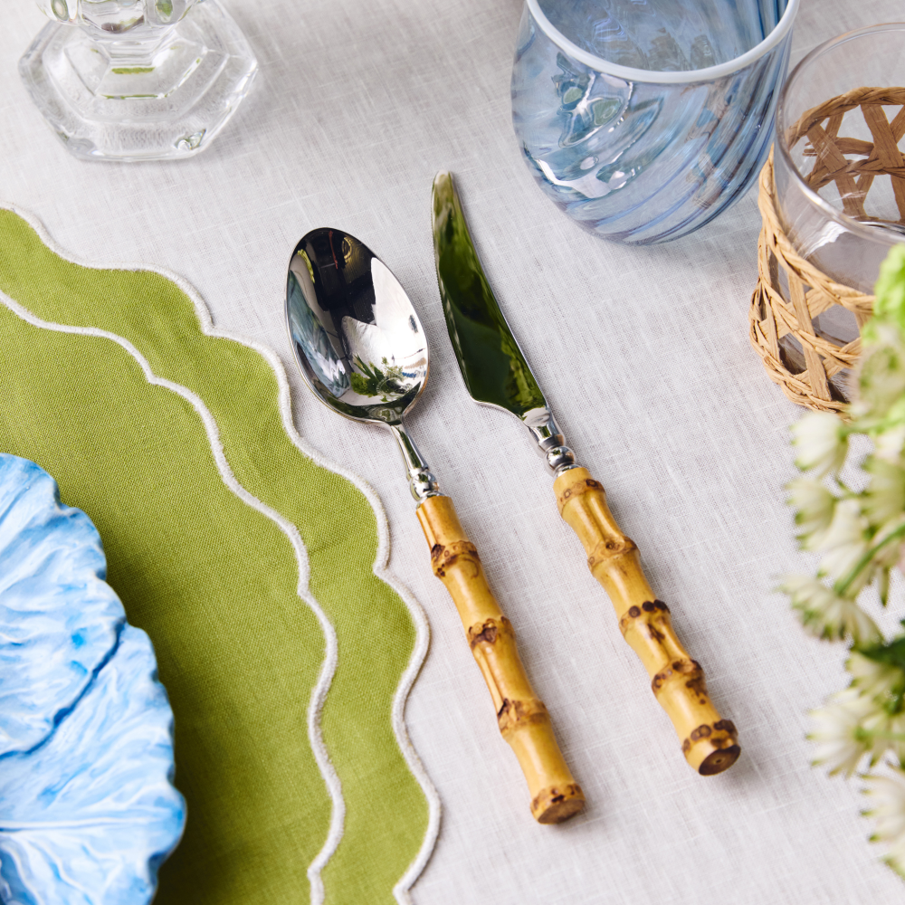 Silver spoon and knife with bamboo handles on a table setting with a green scalloped edge placemat and blue water glass.