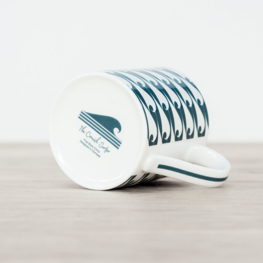 White mug with blue wave design on its side showing The cornish surfer logo on a light grey surface