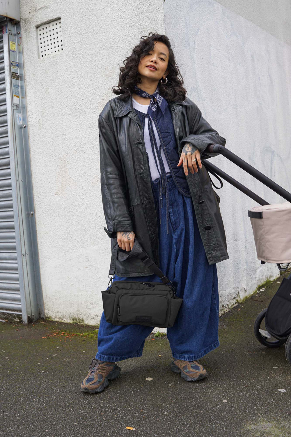Person wearing a long black coat and blue jeans standing against a white wall leaning against a baby buggy holding black bag.