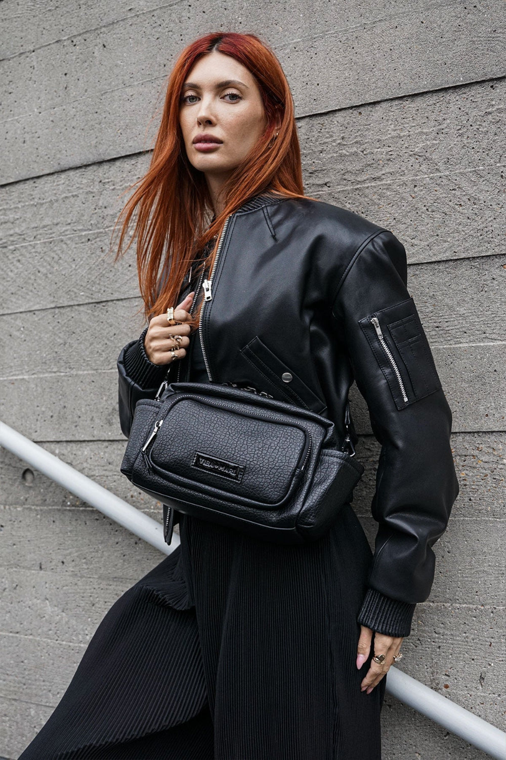 Red haired Woman wearing a black vegan leather jacket and carrying a black crossbody bag against a grey wall.