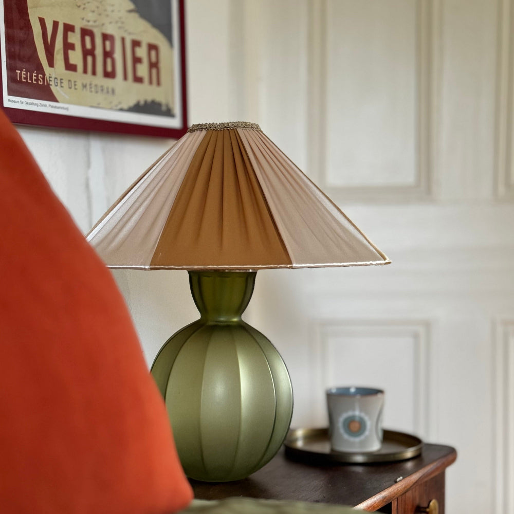 Green lamp with a brown pleated shade on a wooden table, with a blurred red chair and framed poster in the background.