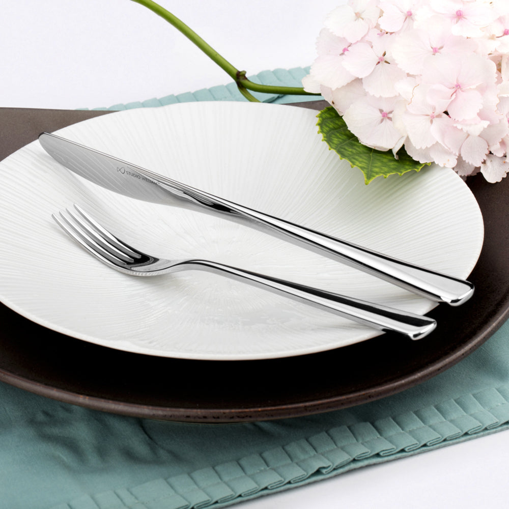 Dinner setting with white plate, black rim, silver knife, and fork on a teal placemat with pink flowers.