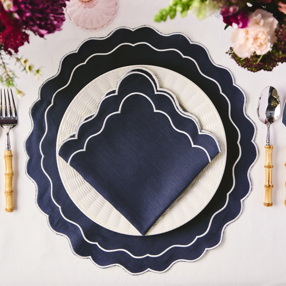 Navy scalloped charger plate with white plate and navy napkin on a table setting with floral decorations.