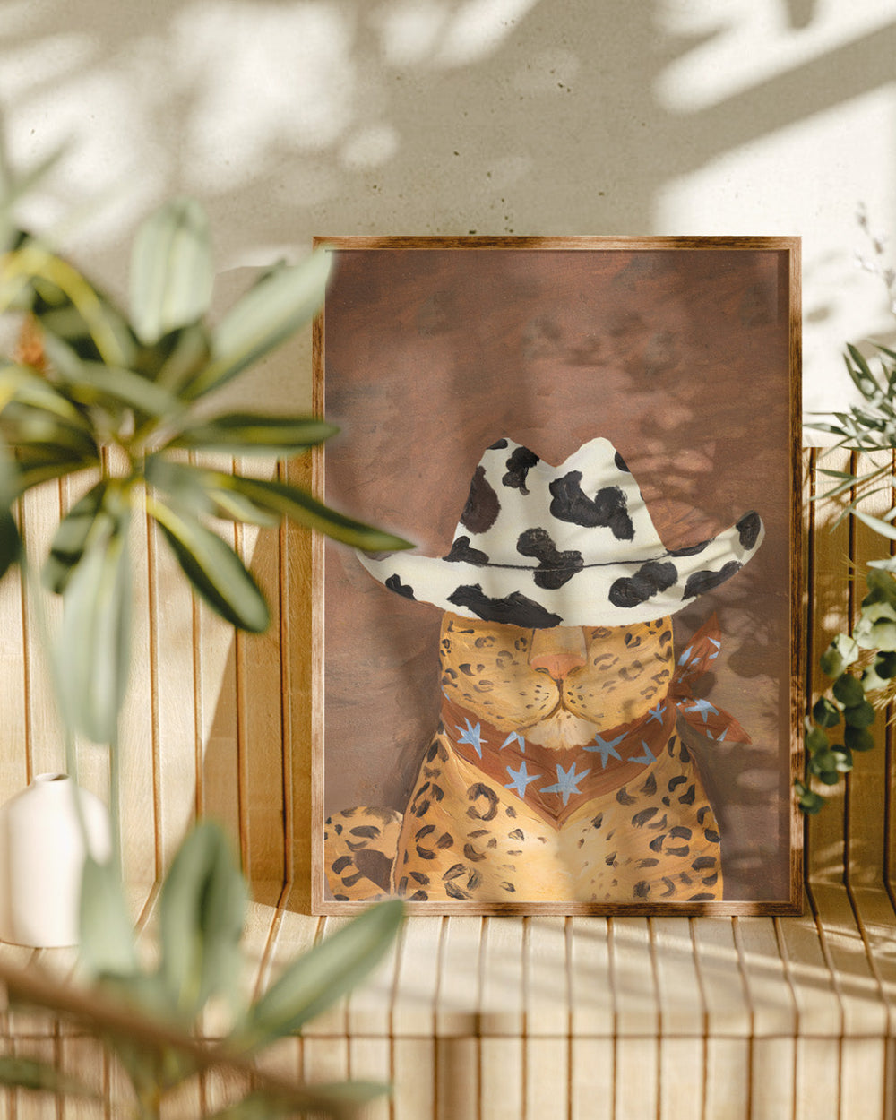Framed artwork of a leopard wearing a cowboy hat with star patterns, placed on a surface with plants in the background.