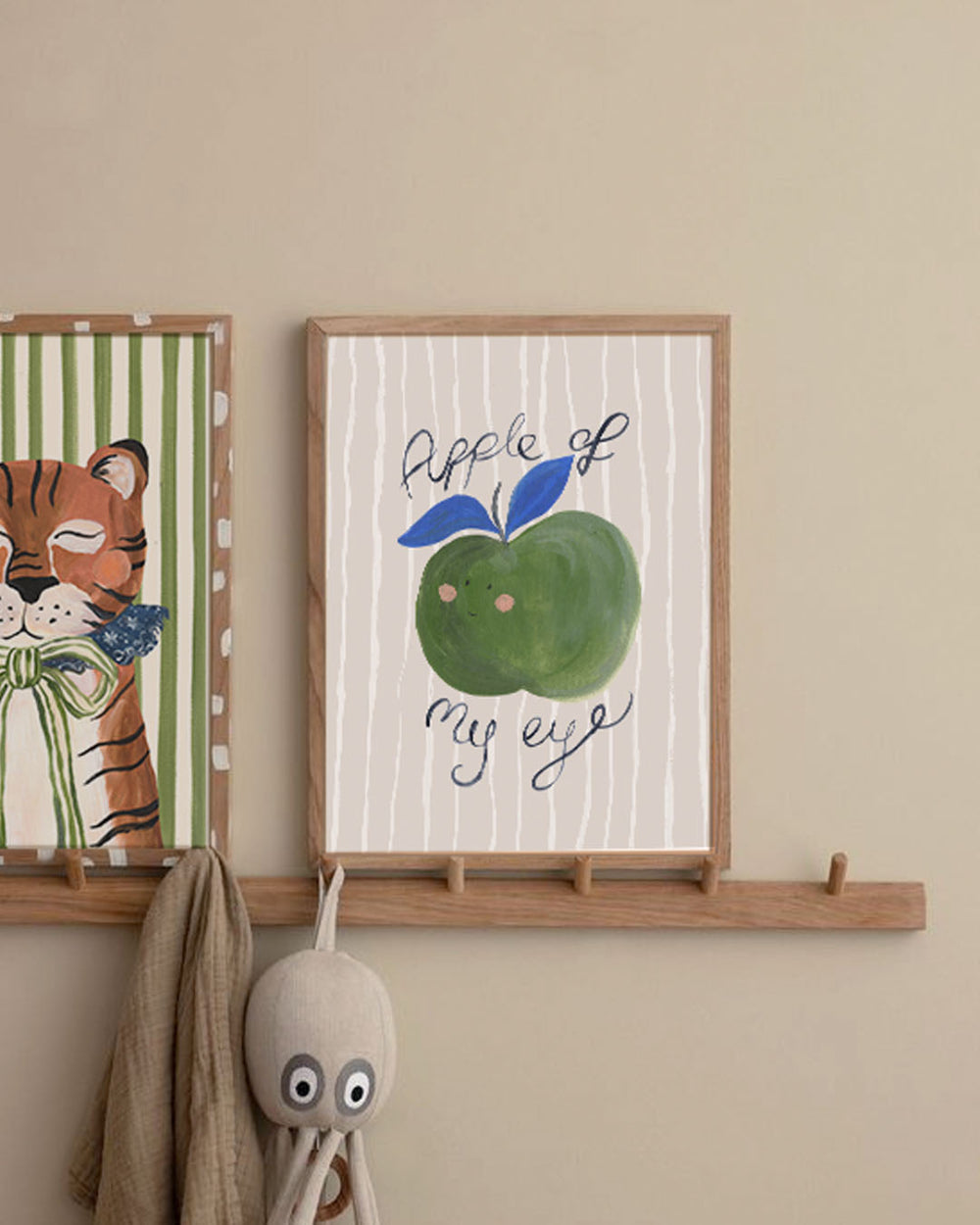 Framed artwork with an apple illustration and text on a wall with a decorative hook and plush toy.