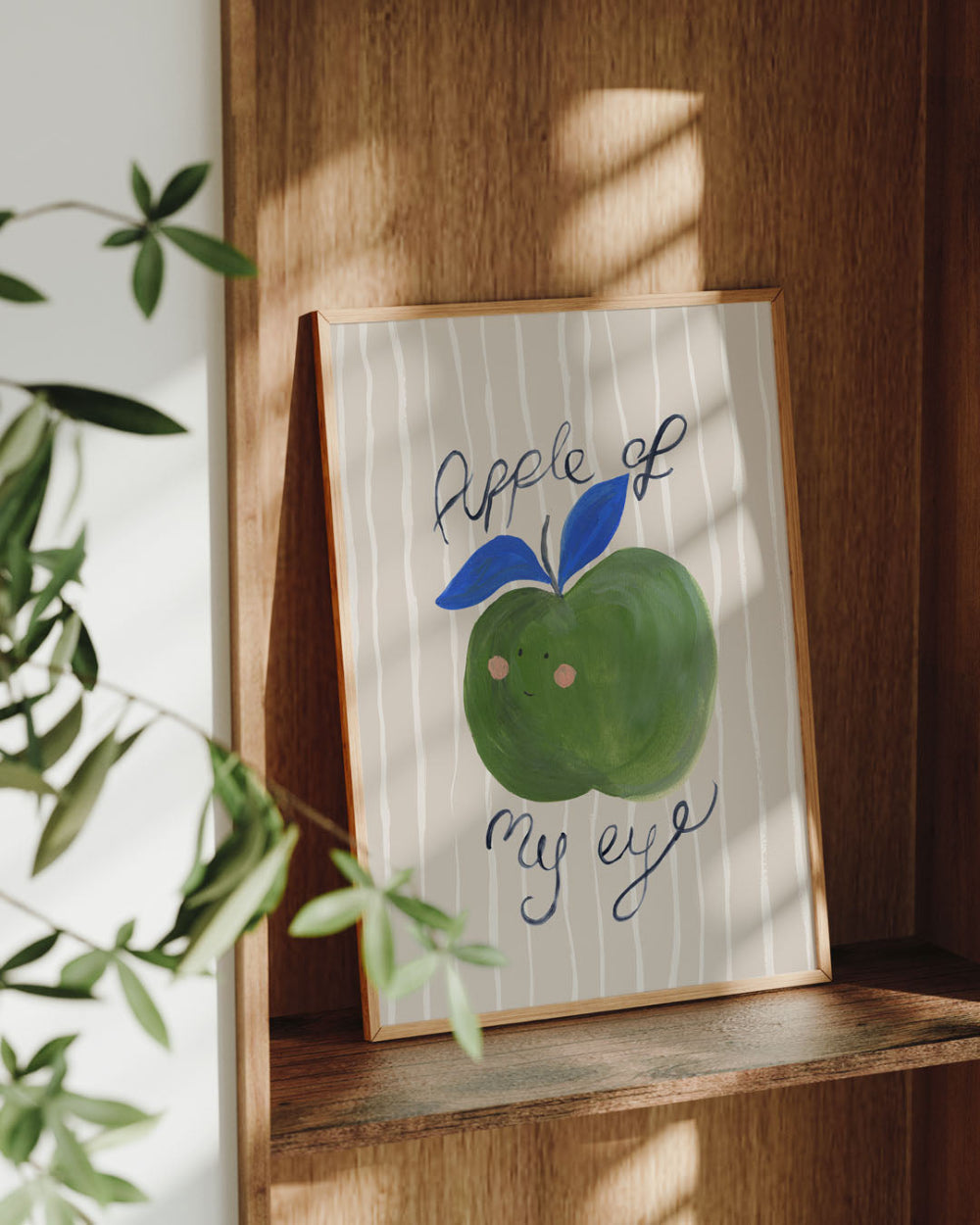 Framed artwork of a green apple with a blue leaf on a wooden shelf.