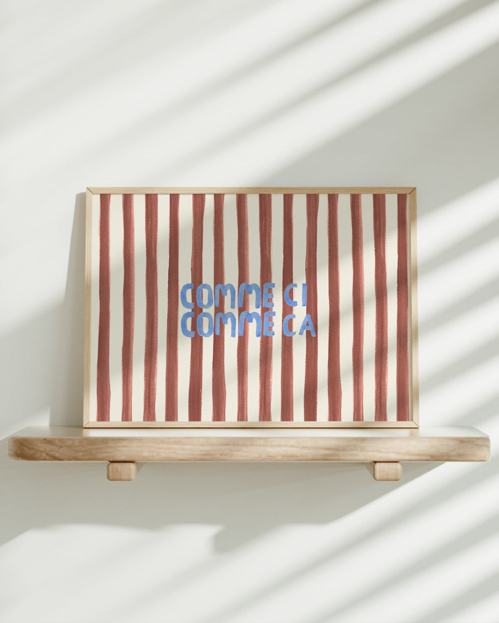 Striped artwork with 'Comme ci, comme ca' text on a wooden shelf against a white wall.