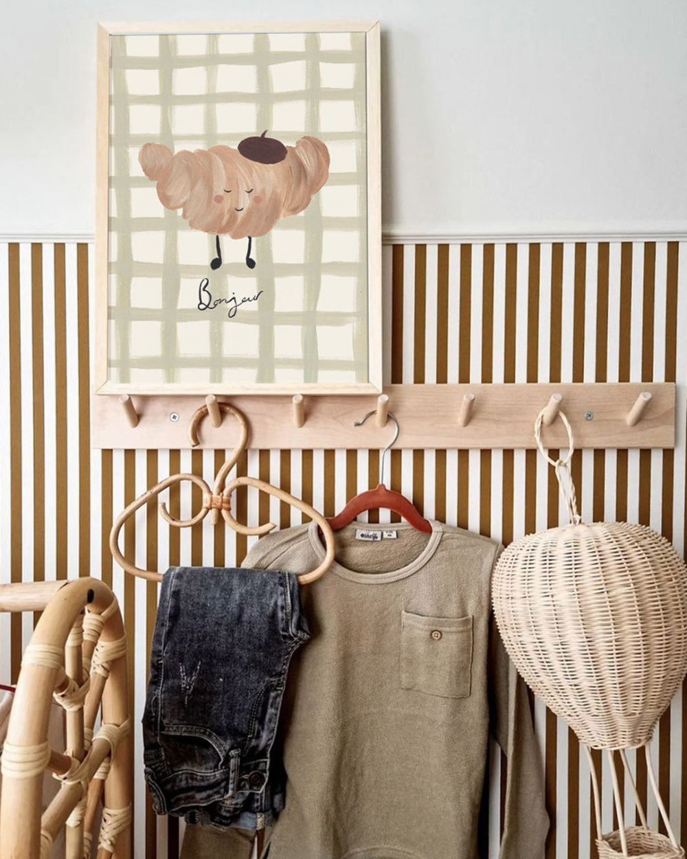 Wall-mounted coat rack with clothes and a decorative croissant childrens painting in a room.
