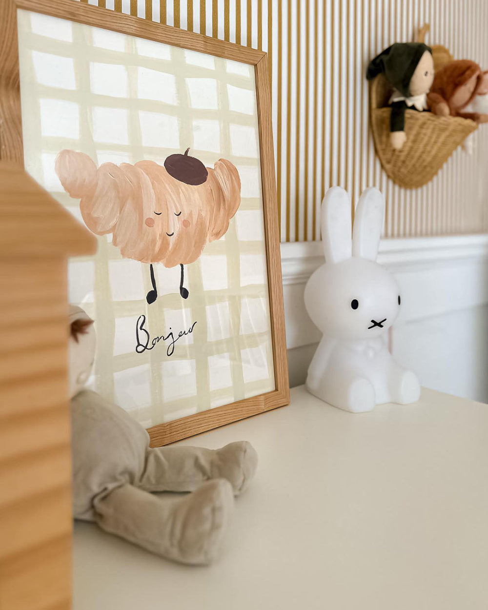 Framed artwork with a croissant cartoon character on a checkered background, placed in a room with toys and decor.