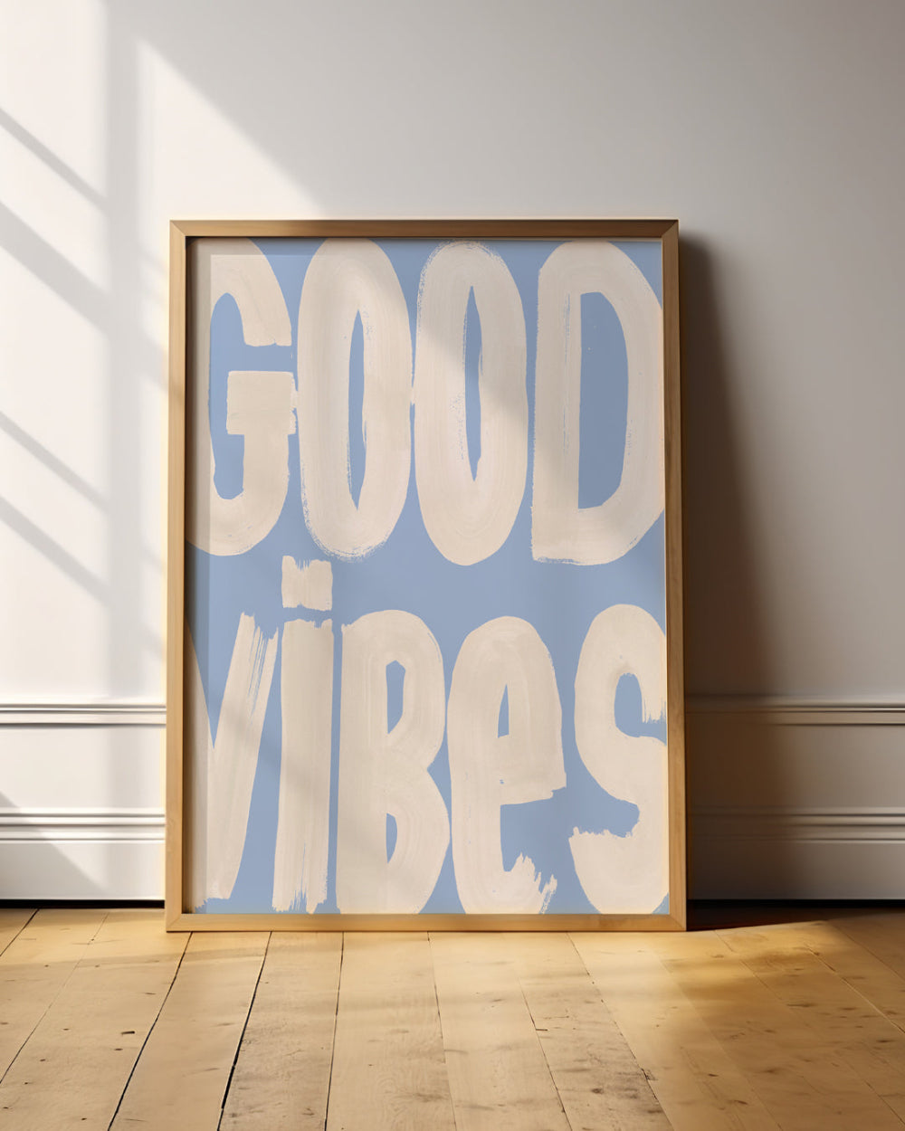 Framed poster with 'GOOD VIBES' text on a light blue background in a room with wooden floor and white walls.