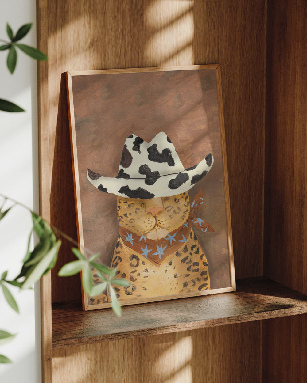 Framed artwork of a cat wearing a cowgirl hat on a wooden shelf.