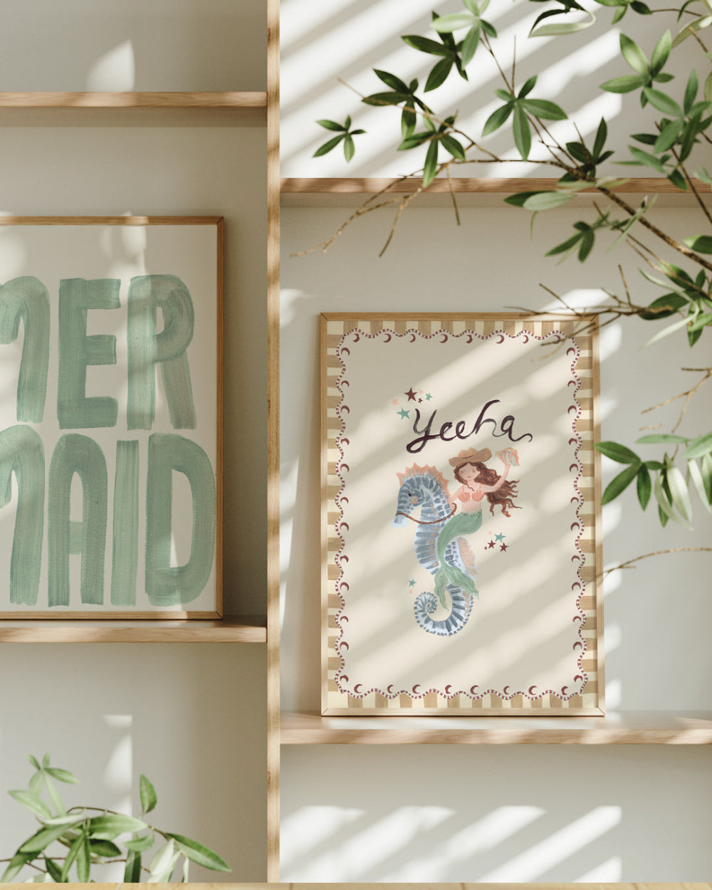 Framed artwork with a mermaid and seahorse design on a shelf with another framed piece in the background.
