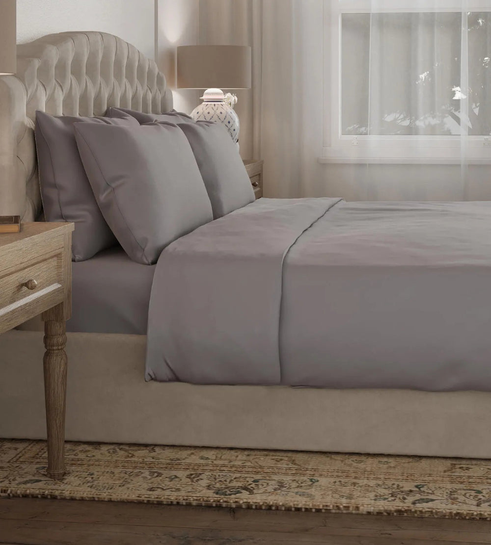 Grey bedding set on a bed in a bedroom with a lamp and window in the background.