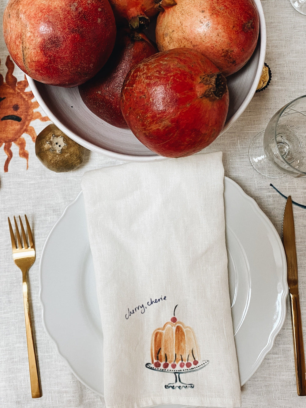 Table setting with pomegranates, a napkin with a cake design, and cutlery on a light surface.