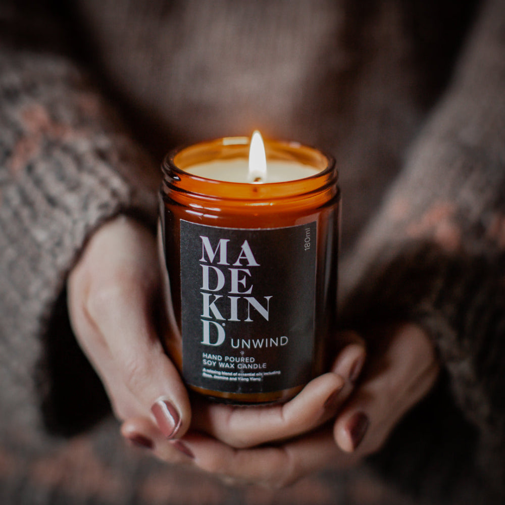 Person holding a lit candle in a jar labeled 'MADEKIND UNWIND' with a blurred background.