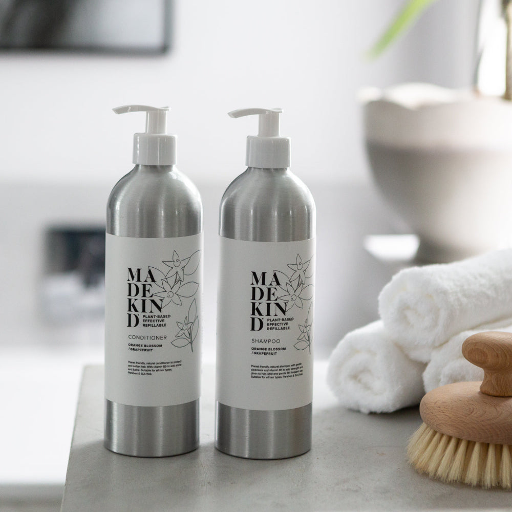 Two bottles of 'MADEKIND' products on a bathroom counter with towels and a brush in the background.