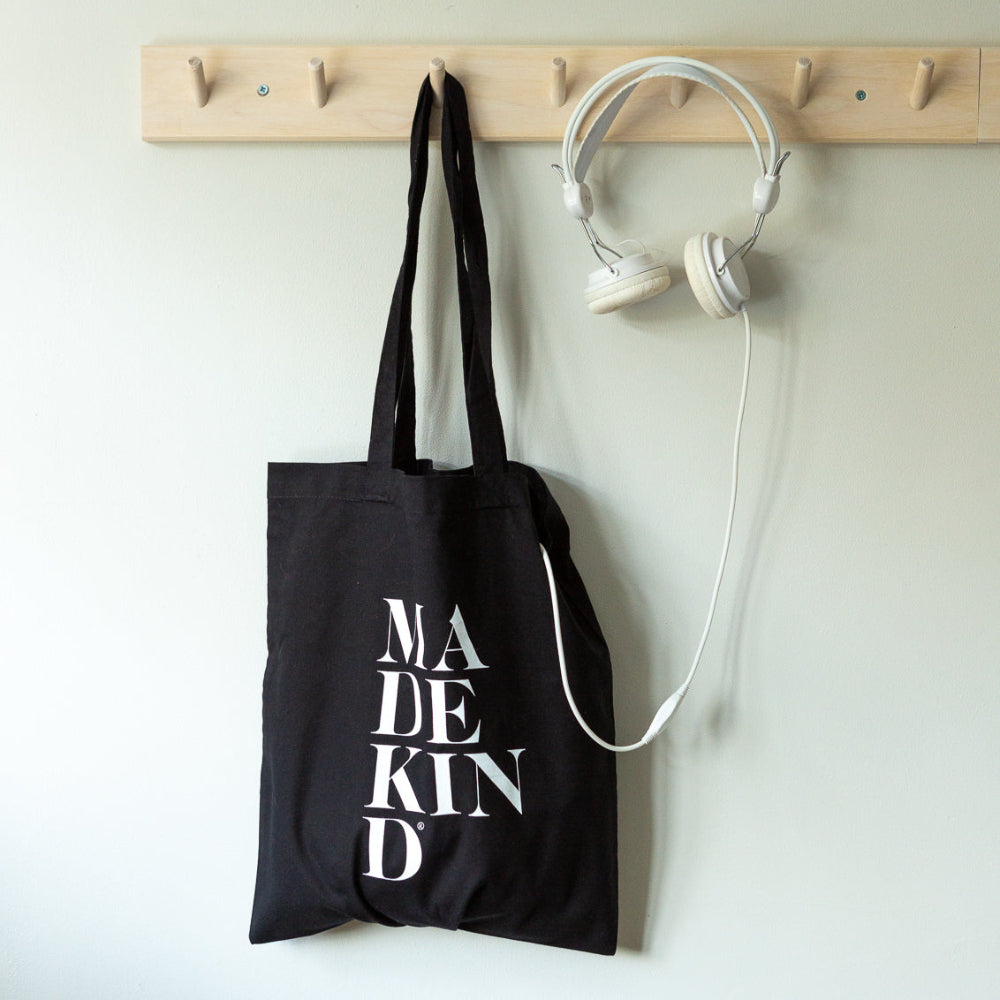 Black tote bag with 'MADE KIND' text hanging on a wooden rack with white headphones.