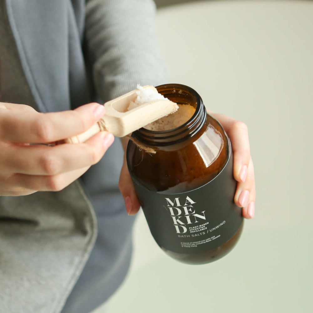 Person holding a brown bath salts jar labelled 'MADEKIND' with a wooden scoop.