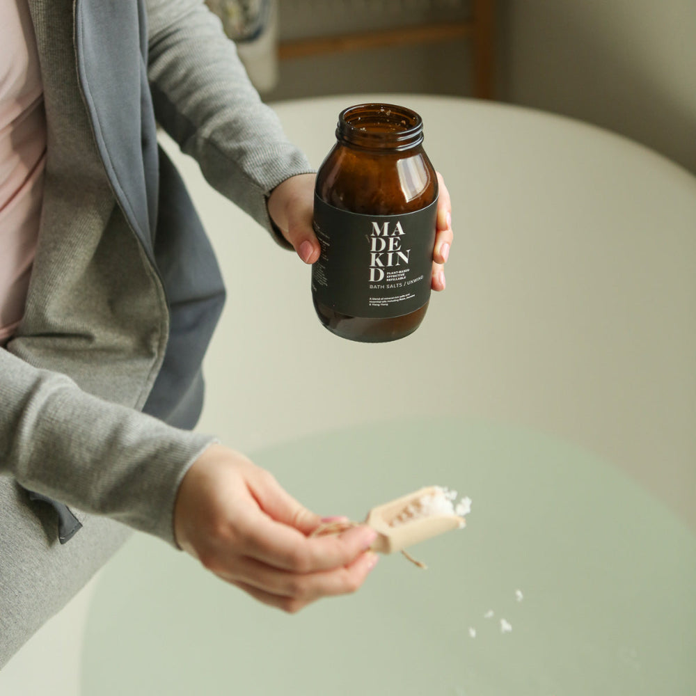 Person holding a brown jar labelled 'MADEKIND' bath salts with a wooden scoop in their other hand over a bathtub.