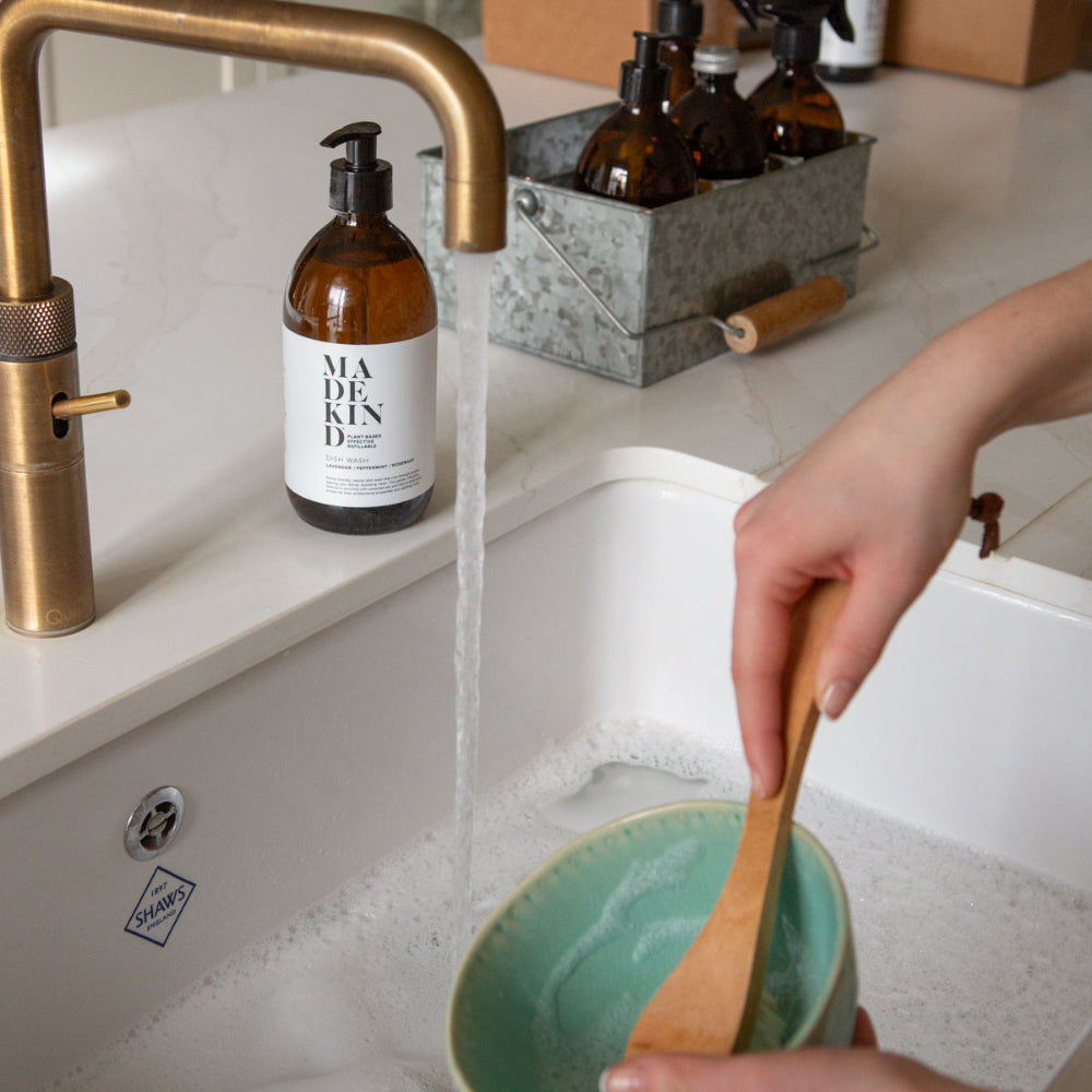 Person washing a green bowl with a wooden spoon in a kitchen sink, with a Madekind Dish wash bottle and other items on the counter.