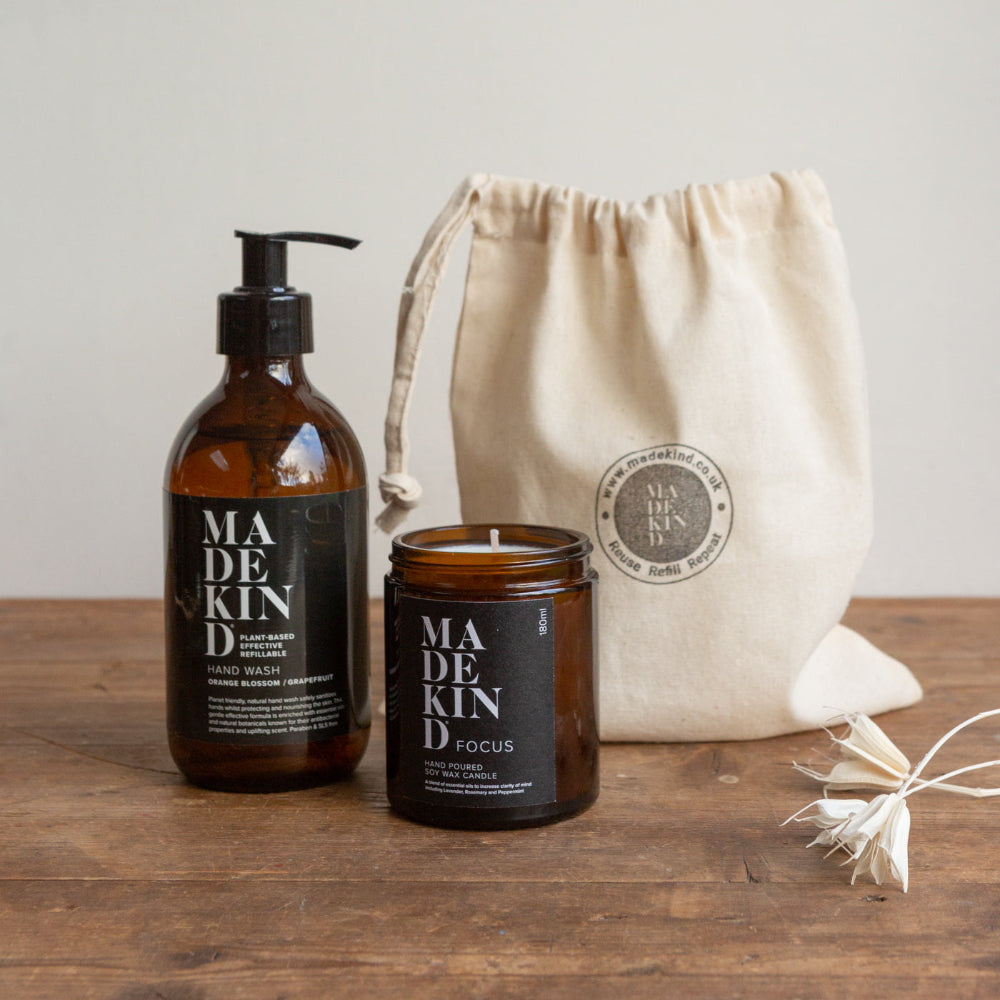 Brown pump bottle hand wash and amber candle jar with 'MADEKIND' branding on a wooden surface with a beige drawstring bag.