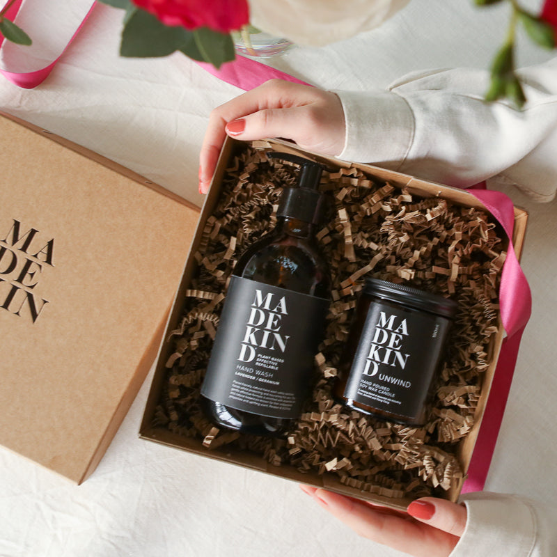 A bottled of 'MADE KIND' ihand eash and unwind candle n a gift box with flowers in the background.