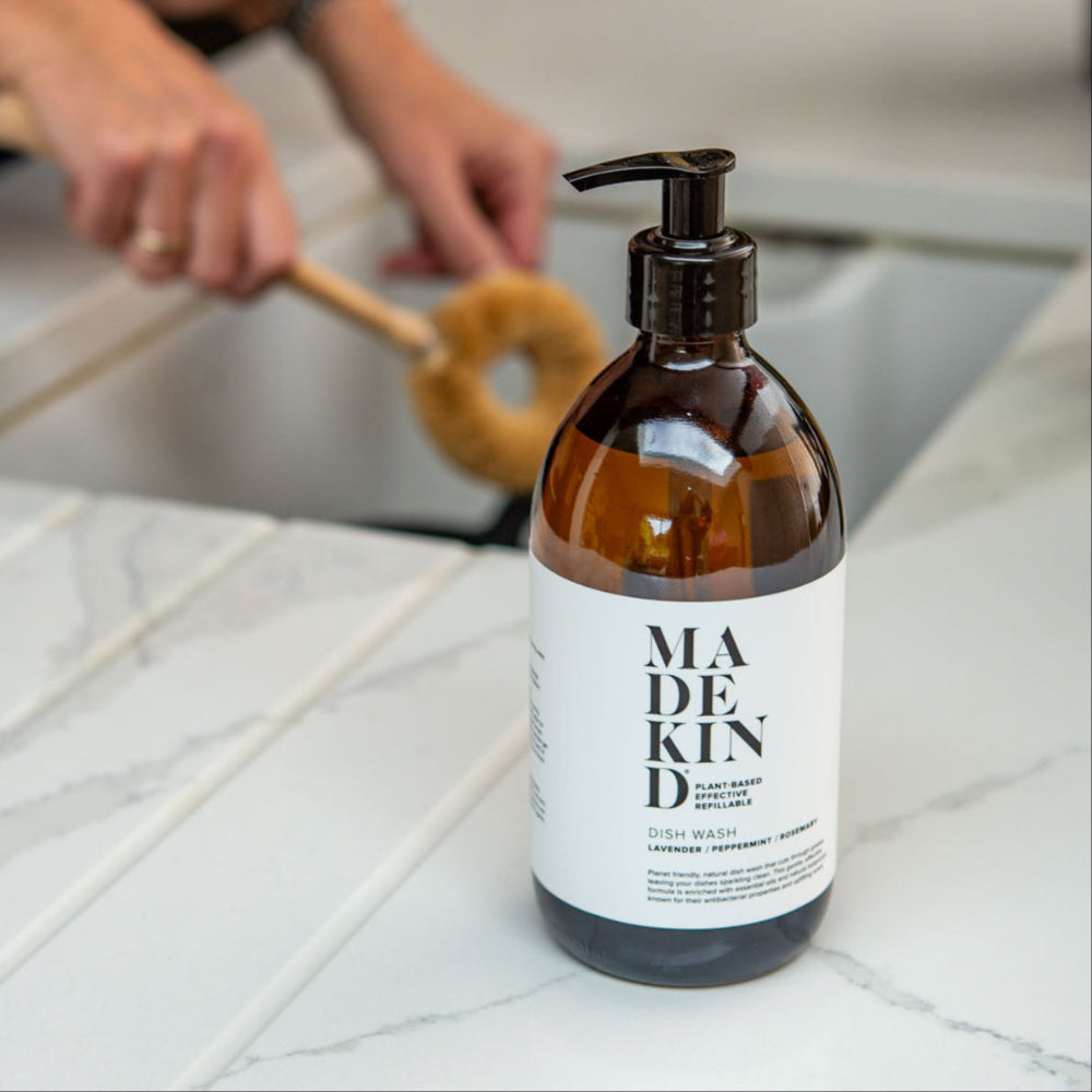 Bottle of Madekind dish wash on a tiled work surface with a person washing dishes in the background.