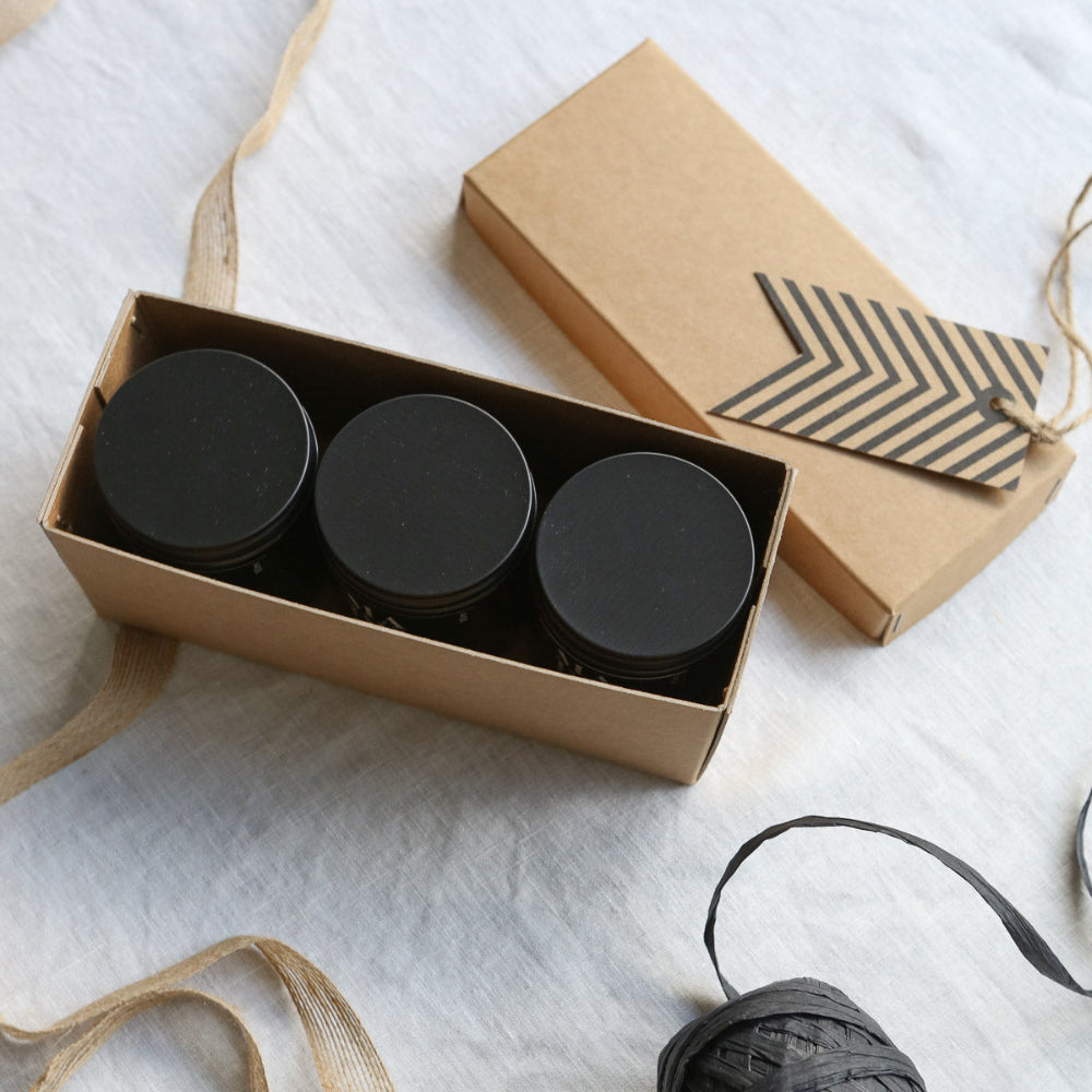 Three black candle jars in a cardboard box with a decorative tag on a white surface.
