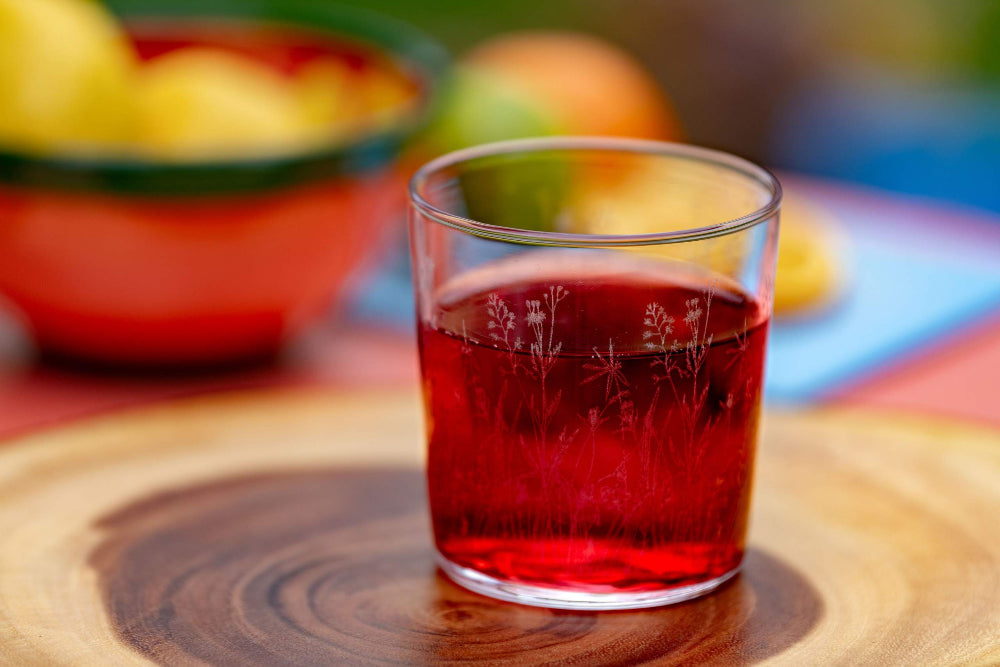 Glass of red liquid on a wooden surface with blurred fruits in the background