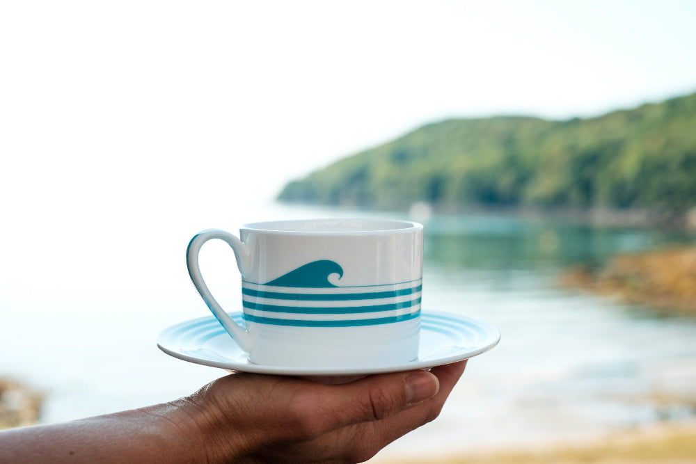 Hand holding a cup and saucer with wave design by a scenic lakeside.