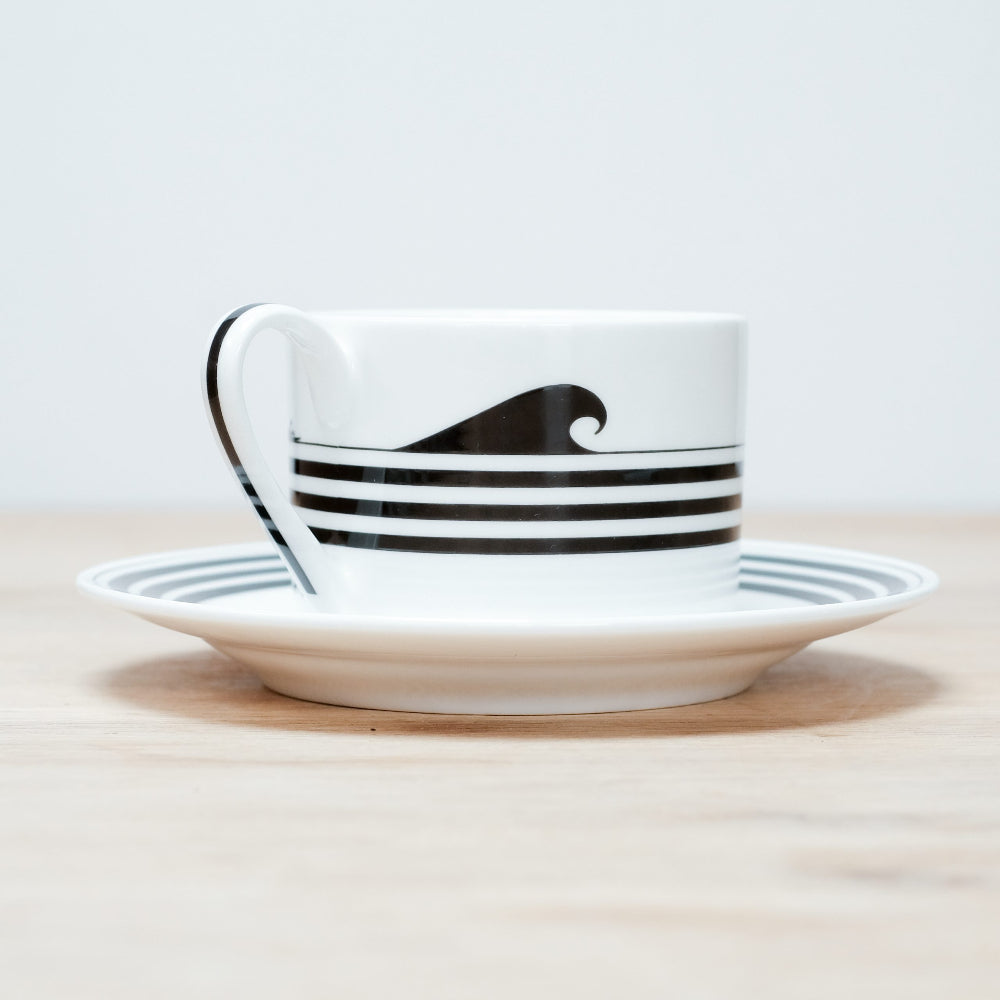 White teacup and saucer with black wave design on a light wooden surface.