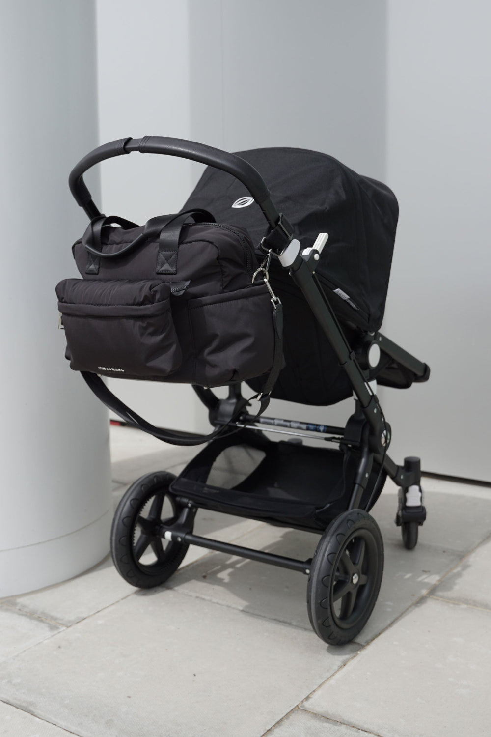 Black stroller with attached bag on a light grey pavement.