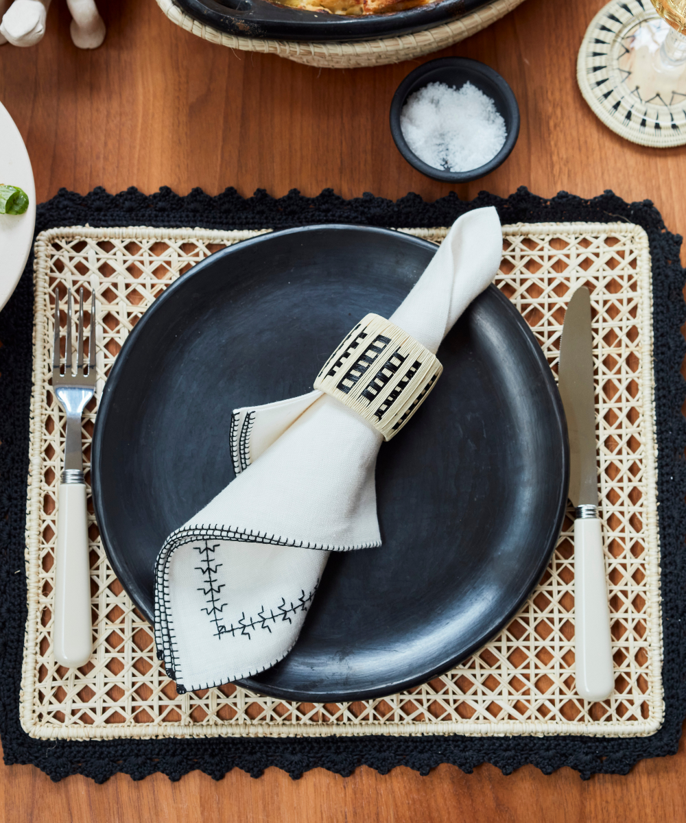 Tablescape setting with white linen napkin and a natural round napkin ring on a black plate and woven placemat.