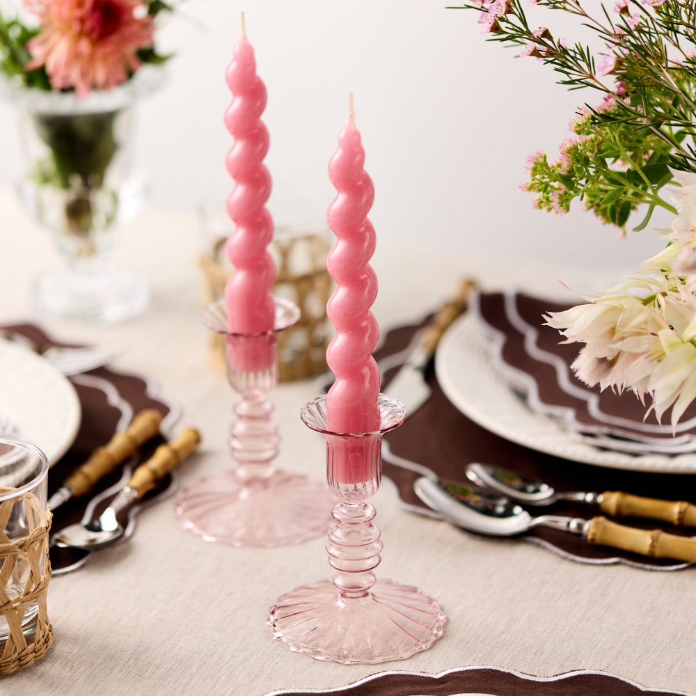 Pink spiral candles in pink glass candle holders on a table setting with flowers and cutlery.