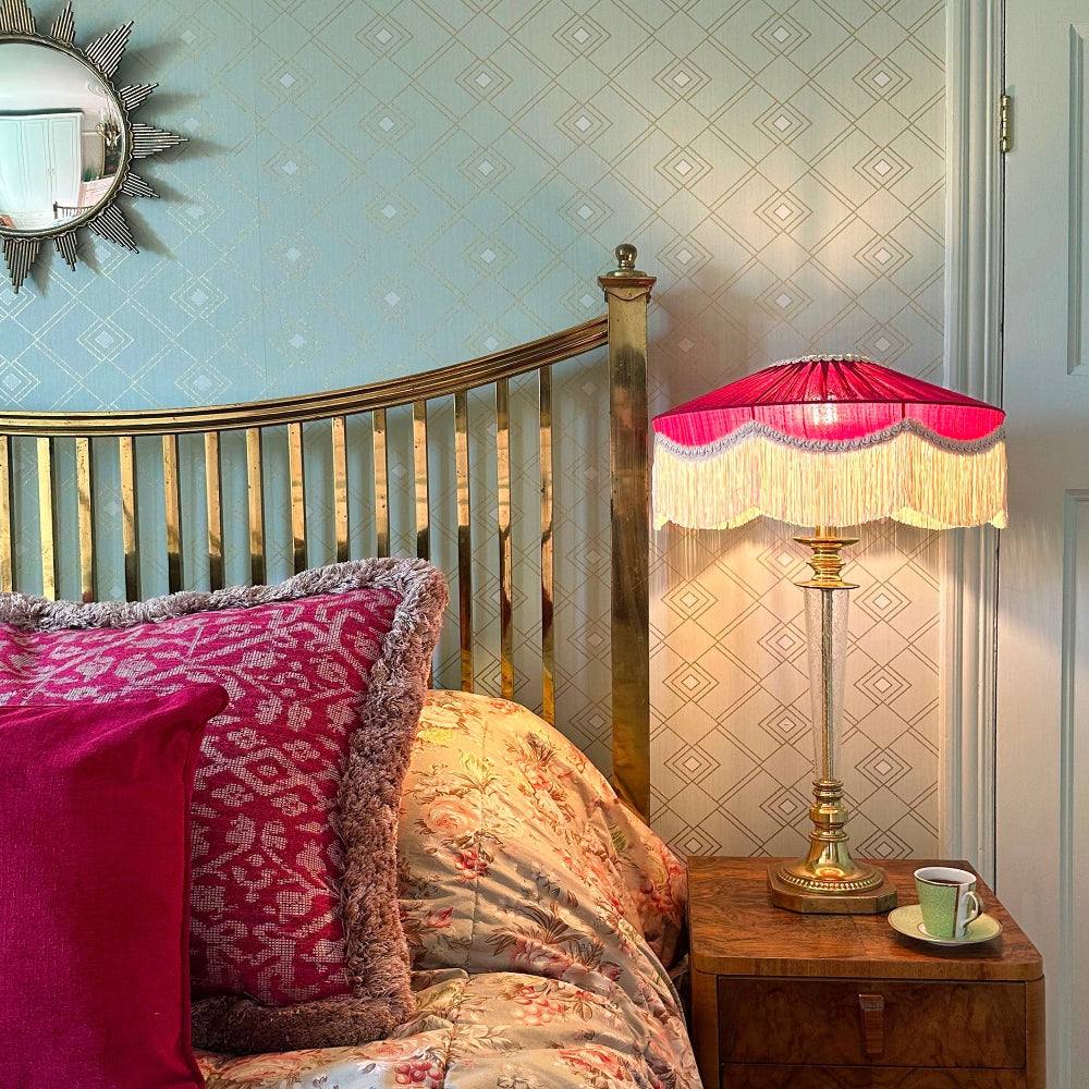 Decorative lamp with pink shade on a wooden side table next to a bed with patterned bedding.