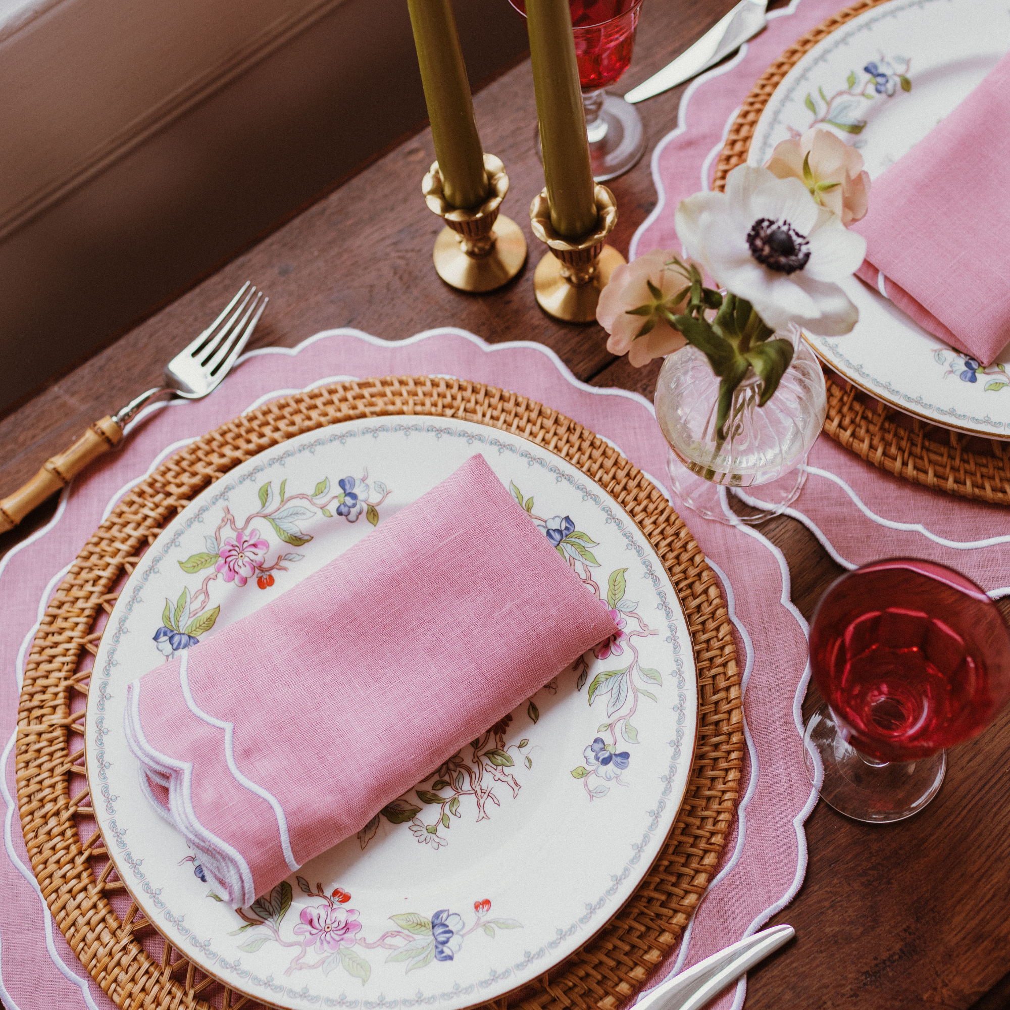 Elegant table setting with pink napkins, floral plates, and red glasses on a wooden table. Pink Scalloped Napkins with white embroidered trim.